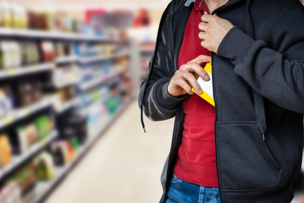 Symbolbild zum Thema Ladendiebstahl im Einzelhandel. Eine Person in einer dunklen Kapuzenjacke steckt im Gang eines Supermarktes heimlich eine Produktverpackung in die Innentasche ihrer Jacke.